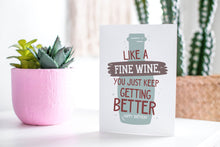 Load image into Gallery viewer, A greeting card featured standing up on a white tabletop with a pink plant pot in the background and some succulents in the pot. There’s a woven basket in the background with a cactus inside. The card features the words &quot;Like a fine wine, you keep getting better, Happy Birthday!” with an illustration of a wine bottle behind the words. 