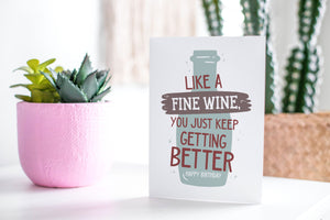 A greeting card featured standing up on a white tabletop with a pink plant pot in the background and some succulents in the pot. There’s a woven basket in the background with a cactus inside. The card features the words "Like a fine wine, you keep getting better, Happy Birthday!” with an illustration of a wine bottle behind the words. 