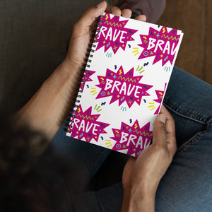 A spiral notebook being held by a person. The white spiral notebook has black spiral binding. The pattern design features the word "brave" inside a hot pink star with sketchy elements around. 