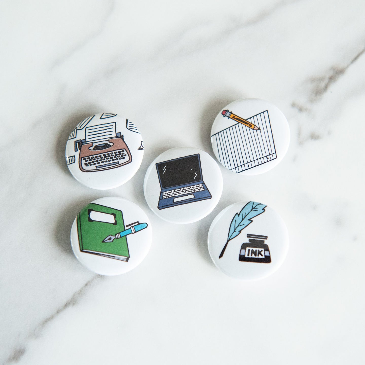 A set of five buttons on a white tabletop. The buttons feature illustrations of a typewriter, laptop, pencil and paper, quill pen, and notebook.