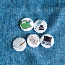 Load image into Gallery viewer, A set of five buttons on a jean jacket. The buttons feature illustrations of a typewriter, laptop, pencil and paper, quill pen, and notebook.