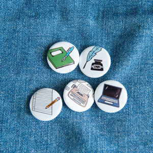 A set of five buttons on a jean jacket. The buttons feature illustrations of a typewriter, laptop, pencil and paper, quill pen, and notebook.