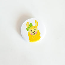 Load image into Gallery viewer, A buttons on a white tabletop. The button features a yellow illustrated llama with green bow on an ear. 