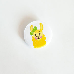 A buttons on a white tabletop. The button features a yellow illustrated llama with green bow on an ear. 