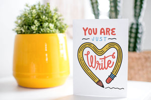 A greeting card is on a table top with a yellow plant pot and a green plant inside. The card features the words “You are just write”with an illustrated pencil in the shape of a heart.