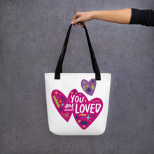 Load image into Gallery viewer, A person holding a white tote bag with black handles. The bag features a design with the words &#39;you are loved&#39; inside two illustrated pink hearts with a smaller, purple heart above. 