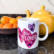 Load image into Gallery viewer, A white mug on a black countertop with a tea pot and oranges behind it. The mug features a design with the words &#39;you are so loved&#39; inside two illustrated pink hearts with a purple heart above it. 