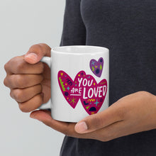 Load image into Gallery viewer, A person holding a white mug. The mug features a design with the words &#39;you are so loved&#39; inside two illustrated pink hearts with a purple heart above it. 