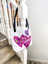 Load image into Gallery viewer, A white tote bag with black handles hanging on a staircase. The bag features a design with the words &#39;you are loved&#39; inside two illustrated pink hearts with a smaller, purple heart above. 