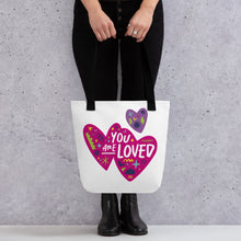 Load image into Gallery viewer, A person holding a white tote bag with black handles. The bag features a design with the words &#39;you are loved&#39; inside two illustrated pink hearts with a smaller, purple heart above. 