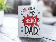 Load image into Gallery viewer, A greeting card featured on a black, wood coffee table. There’s a white planter in the background with a green plant. There’s also a gray sofa in the background with a white pillow. The card features the words &quot;You are my hero Dad.” 