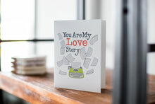 Load image into Gallery viewer, A card on a wood tabletop with an object in the background that is out of focus. The card features the words “You are my love story” with an illustrated typewriter and scattered papers.