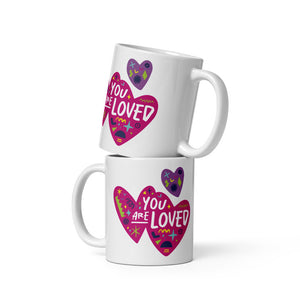 Two white mugs stacked on top of each other. The mug features a design with the words 'you are so loved' inside two illustrated pink hearts with a purple heart above it. 