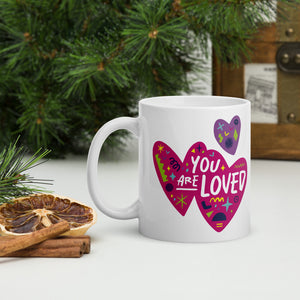 A white mug on a tabletop with part of a pine tree in the background. The mug features a design with the words 'you are so loved' inside two illustrated pink hearts with a purple heart above it. 