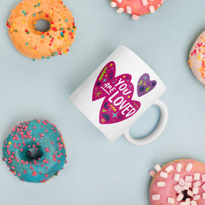 A white mug laying on a tabletop with doughnuts around it. The mug features a design with the words 'you are so loved' inside two illustrated pink hearts with a purple heart above it. 