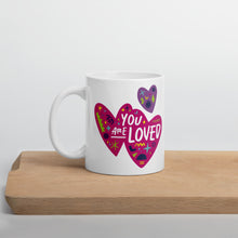 Load image into Gallery viewer, A white mug on a wood board. The mug features a design with the words &#39;you are so loved&#39; inside two illustrated pink hearts with a purple heart above it. 