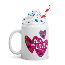 Load image into Gallery viewer, A white mug on a white background with whipped cream and a straw. The mug features a design with the words &#39;you are so loved&#39; inside two illustrated pink hearts with a purple heart above it. 