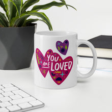 Load image into Gallery viewer, A white mug on a desktop. The mug features a design with the words &#39;you are so loved&#39; inside two illustrated pink hearts with a purple heart above it. 