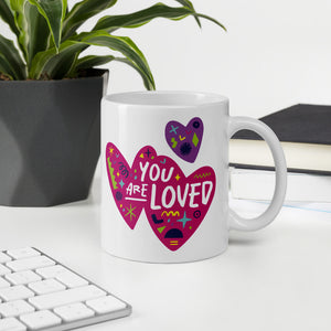 A white mug on a desktop. The mug features a design with the words 'you are so loved' inside two illustrated pink hearts with a purple heart above it. 