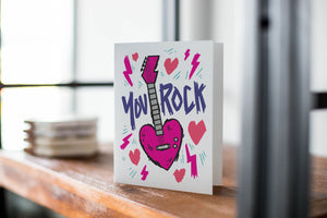A card on a wood tabletop with an object in the background that is out of focus. The card features the words “You rock” with an illustrated heart shaped guitar. 