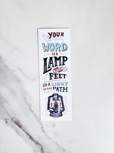A bookmark laying on a marble tabletop. The bookmark has a white background with the words "Your word is a lamp to my feet and a light to my path" and an illustrated lamp on the bottom. 