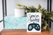 Load image into Gallery viewer, A greeting card is on a table top with a present in blue wrapping paper in the background. On top of the present is a candle and some greenery from a plant too. The card features the words “You’ve got game” with an illustrated gaming controller. 
