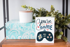 A greeting card is on a table top with a present in blue wrapping paper in the background. On top of the present is a candle and some greenery from a plant too. The card features the words “You’ve got game” with an illustrated gaming controller. 