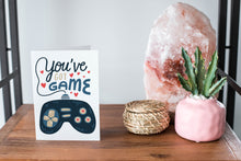 Load image into Gallery viewer, A greeting card featured standing up on a white tabletop with a pink plant pot in the background and some succulents in the pot. There’s a woven basket in the background with a cactus inside. The card features the words “You’ve got game” with an illustrated gaming controller.