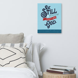 An art canvas hanging on a white wall above a bed with white pillows and cushions. Next to the bed is a pile of books on a wooden side table. The print is bright blue with the verse 'Be Still and Know that I am God' illustrated in a bold typographic style.