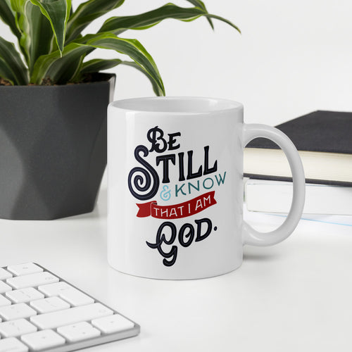 A white coffee mug stands on a white desk next to a computer keyboard and a houseplant. The mug features the verse 'Be Still and Know that I am God' illustrated in a bold typographic style, with black lettering and a red accent.