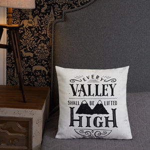 A monochrome square pillow sits on a grey bed with an upholstered headboard. The pillow design is black on a white background, and reads 'Every valley shall be lifted high' in a variety of typographic lettering, with flourishes and an illustration of two mountain peaks. Next to the bed is a side table with a lamp.