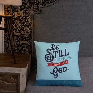 A bright blue cushion sits on a dark grey bed next to a bedside table and small lamp. The cushion features the verse 'Be Still and Know that I am God' illustrated in a bold typographic style.