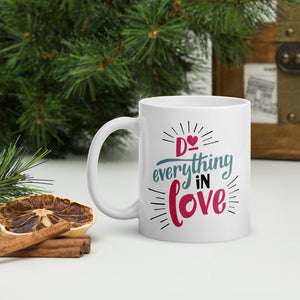 A white mug sits on a white table next to dried fruit and cinnamon sticks and evergreen foliage. The mug reads "Do everything in love' in bright pink and blue lettering, with black dashes coming out from the words.