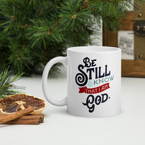 A white coffee mug stands on a white desk surrounded by Christmas foliage, dried oranges and cinnamon sticks. The mug features the verse 'Be Still and Know that I am God' illustrated in a bold typographic style, with black lettering and a red accent.