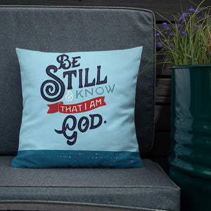 A bright blue cushion sits on an outdoor sofa next to a planter filled with purple flowers. The cushion features the verse 'Be Still and Know that I am God' illustrated in a bold typographic style.