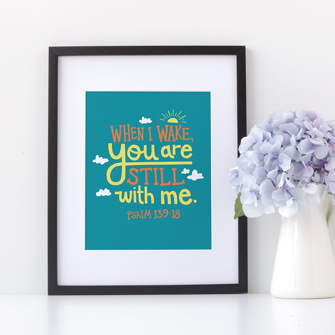 A small turquoise print in a black frame sits on a white shelf. The print reads 'When I wake you are still with me, Psalm 139:18' in orange and yellow lettering, illustrated with a small yellow sun and little grey clouds. Beside the print is a small white jug of blue flowers.