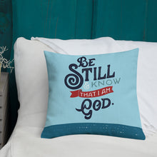 Load image into Gallery viewer, A bright blue cushion sits on a white bed against a teal wall. The cushion features the verse &#39;Be Still and Know that I am God&#39; illustrated in a bold typographic style.
