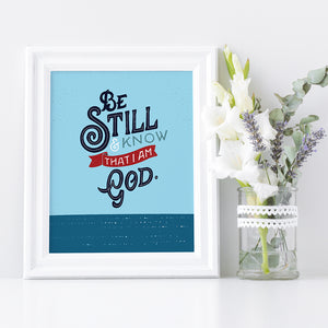 A small white framed print sits next a to a small vase of flowers. The print is bright blue with the verse 'Be Still and Know that I am God' illustrated in a bold typographic style.