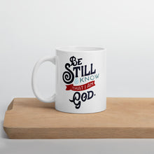 Load image into Gallery viewer, A white coffee mug stands on a light wooden shelf. The mug features the verse &#39;Be Still and Know that I am God&#39; illustrated in a bold typographic style, with black lettering and a red accent.