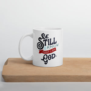 A white coffee mug stands on a light wooden shelf. The mug features the verse 'Be Still and Know that I am God' illustrated in a bold typographic style, with black lettering and a red accent.