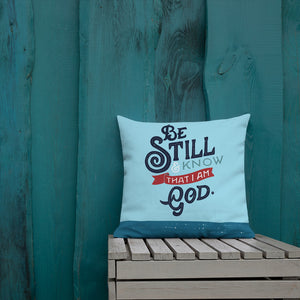 A bright blue cushion sits on a slatted garden seat against a teal fence. The cushion features the verse 'Be Still and Know that I am God' illustrated in a bold typographic style.