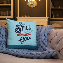 Load image into Gallery viewer, A bright blue cushion sits on a chunky throw on a grey upholstered sofa. The cushion features the verse &#39;Be Still and Know that I am God&#39; illustrated in a bold typographic style.