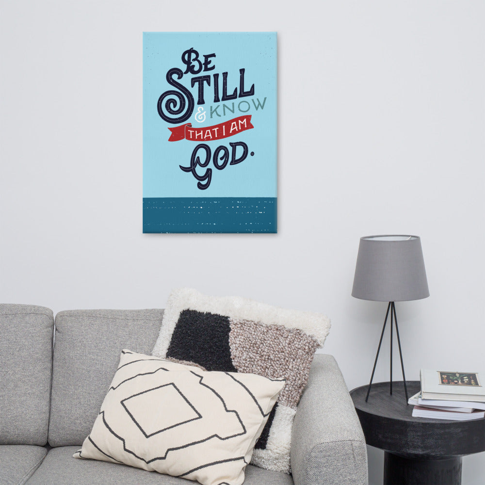 An art canvas hanging on a white wall above a grey sofa with black and white cushions. The print is bright blue with the verse 'Be Still and Know that I am God' illustrated in a bold typographic style.