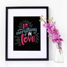 Load image into Gallery viewer, A small black art print in a black frame leans against a white wall. Beside it is a vase of pink flowers. The print reads &quot;Do everything in love&quot; in bright pink and blue hand-lettering style, with white dashes around the words.