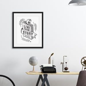A black frame above a desk with artwork printed on white paper. The artwork features hand drawn lettering with the words "Live Your Story" inside an illustrated book. 