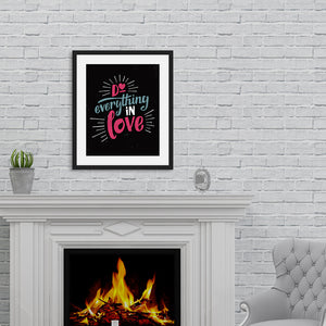 A black art print in a black frame hangs on a painted brick wall, above a lit fireplace. The print reads "Do everything in love" in bright pink and blue hand-lettering style, with white dashes around the words.