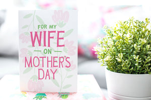 A greeting card is featured on a desktop with a green plant in the background. The card features illustrated flowers in the background with the words “For my wife on Mother’s Day.”