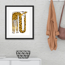 Load image into Gallery viewer, A black frame on a wall with a yellow giraffe illustration. The frame is on the wall above a white side table and next to a coat rack. 
