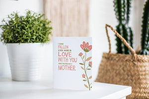 A greeting card is featured on a white tabletop with a white planter in the background with a green plant. There’s a woven basket in the background with a cactus inside. The card features illustrated flowers on the right hand side of the card. The petals are muted pink and the stem and leaves are green and light brown. To the left of the flowers the card reads "I have fallen more in love with you since you became a mother." 