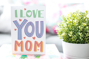A greeting card is featured on a desktop with a green plant to the side. The card features a couple illustrated hearts with the words “I love you mom.”